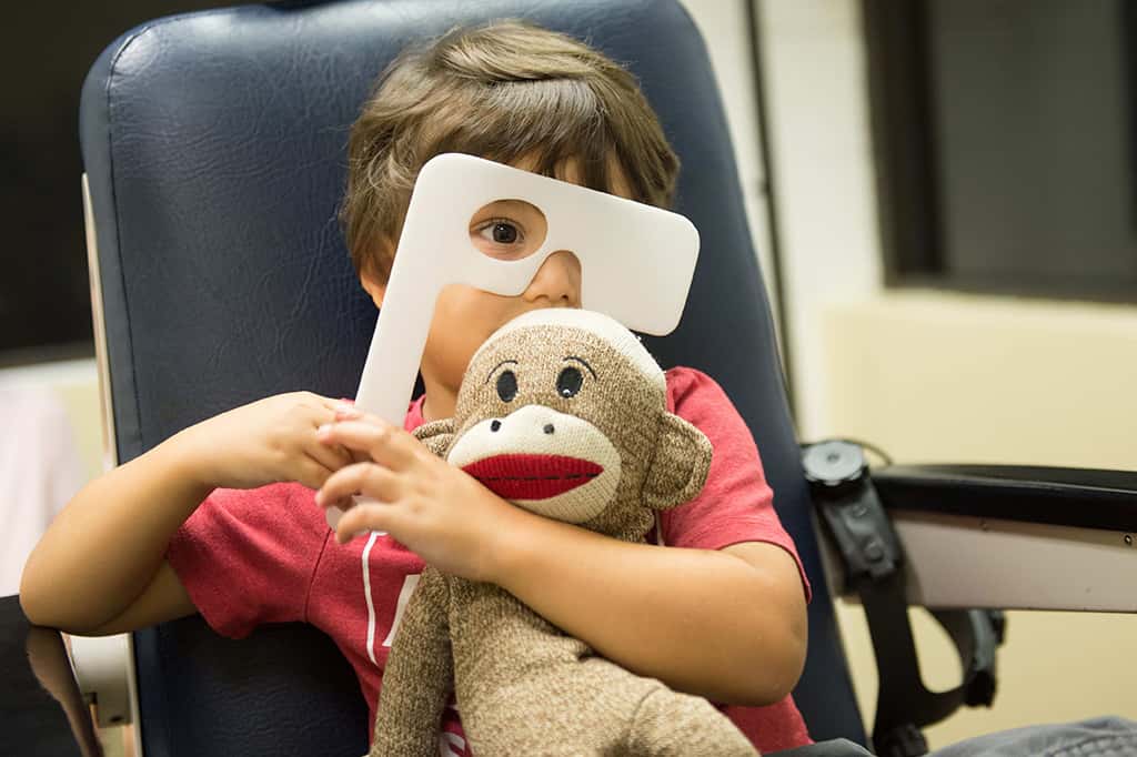 Young boy in red shirt holding a stuffed animal taking an eye test for free at SEE Vision Care